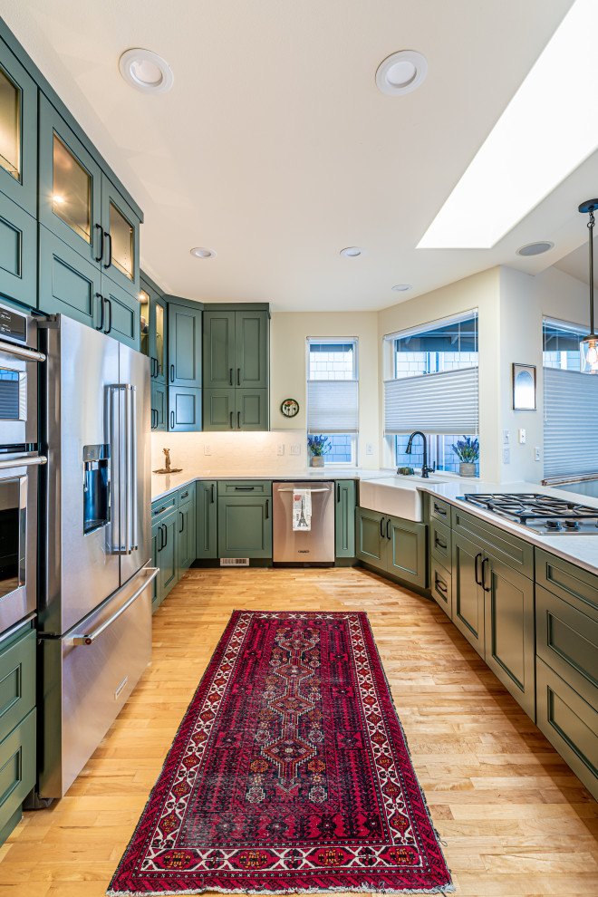 Mid-sized transitional u-shaped medium tone wood floor and brown floor open concept kitchen photo in Seattle with a farmhouse sink, green cabinets, white backsplash, stainless steel appliances, white countertops, flat-panel cabinets, quartz countertops, ceramic backsplash and a peninsula