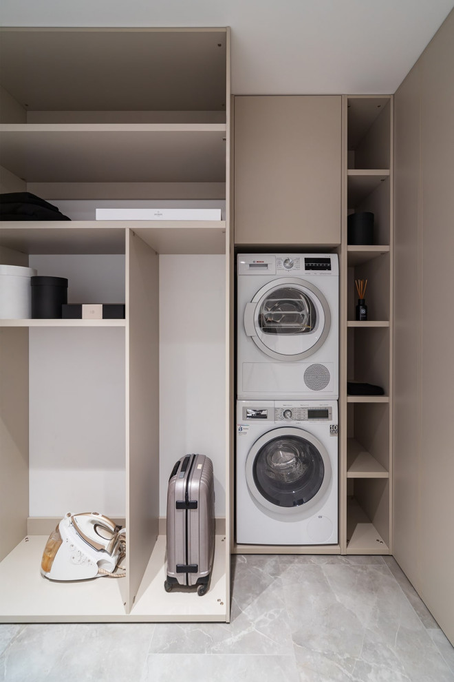 This is an example of a small contemporary l-shaped dedicated laundry room in Yekaterinburg with a drop-in sink, flat-panel cabinets, beige cabinets, quartz benchtops, white splashback, engineered quartz splashback, grey walls, porcelain floors, a stacked washer and dryer, grey floor and white benchtop.
