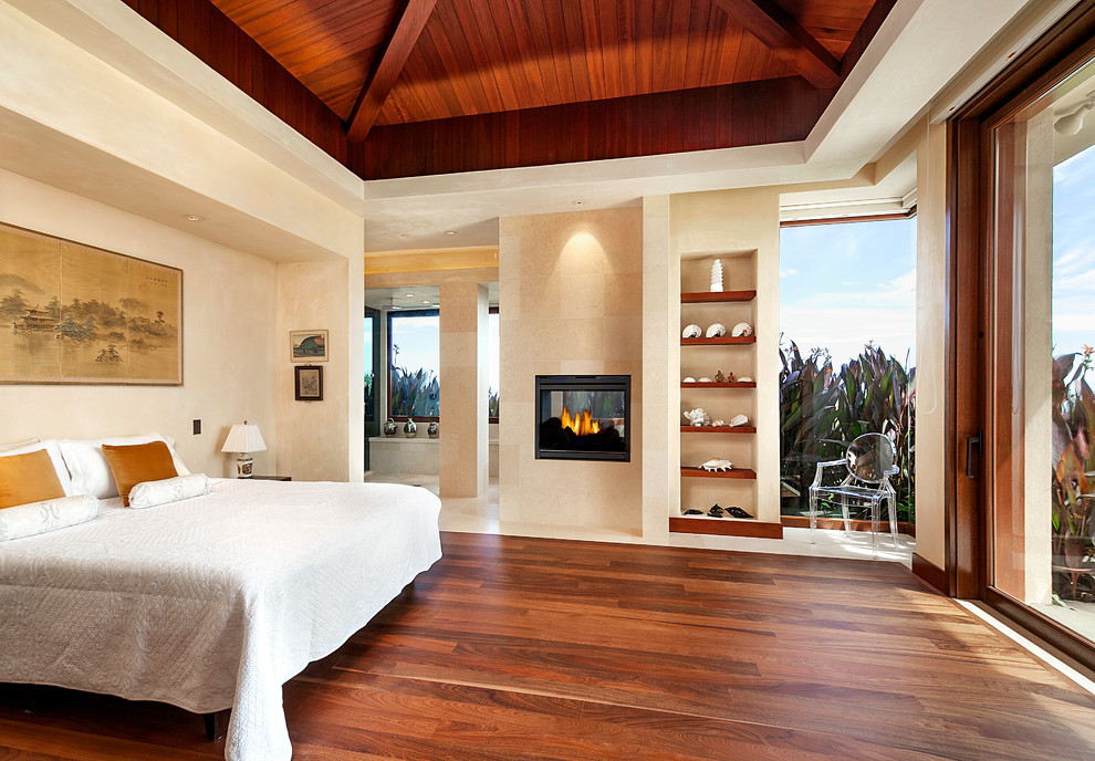 This is an example of a tropical master bedroom in Santa Barbara with beige walls, dark hardwood floors and a two-sided fireplace.