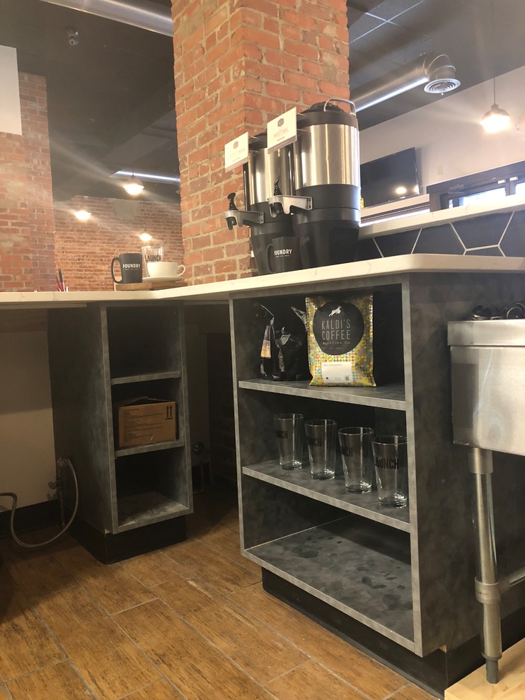 This is an example of a large industrial l-shaped seated home bar in Omaha with a drop-in sink, open cabinets, grey cabinets, onyx benchtops, black splashback, ceramic splashback, ceramic floors, brown floor and white benchtop.