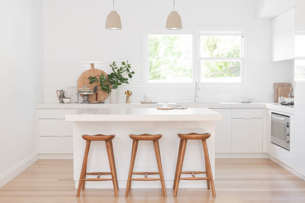 Contemporary l-shaped kitchen in Geelong with an undermount sink, flat-panel cabinets, white cabinets, white splashback, stainless steel appliances, light hardwood floors, with island, beige floor and white benchtop.
