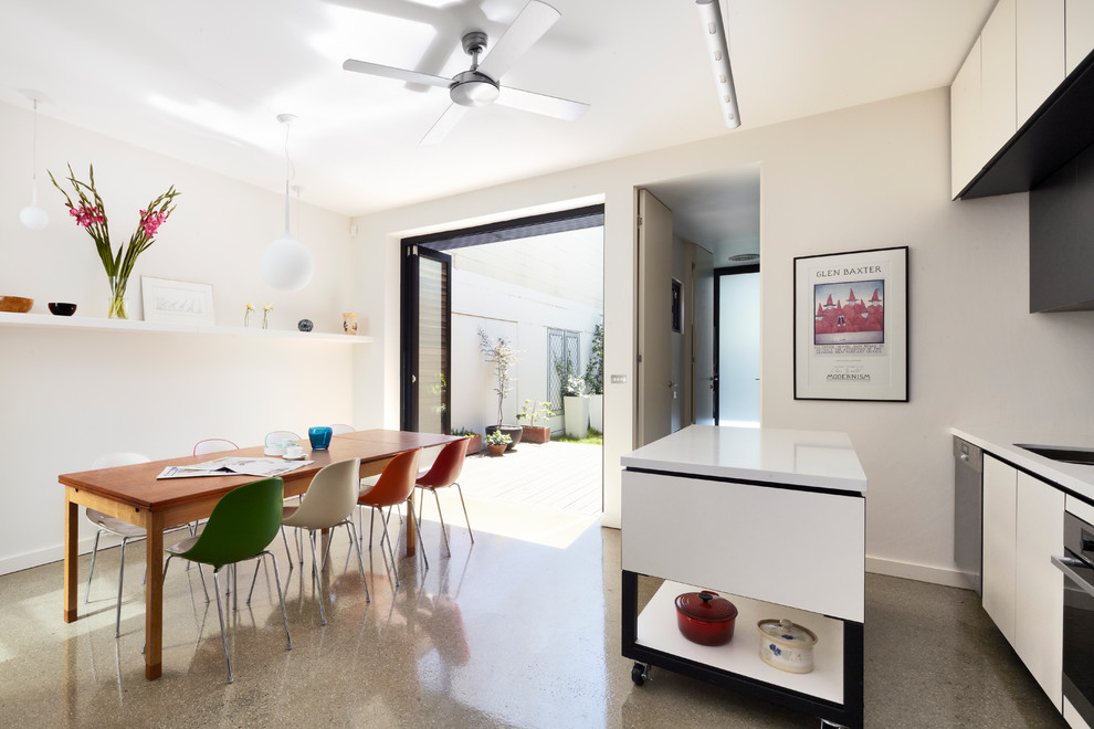 Inspiration for a mid-sized contemporary single-wall eat-in kitchen in Melbourne with a drop-in sink, flat-panel cabinets, white cabinets, black splashback, glass sheet splashback, stainless steel appliances, concrete floors, with island, brown floor and white benchtop.