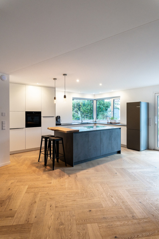 Photo of a large contemporary l-shaped open plan kitchen in Frankfurt with an integrated sink, flat-panel cabinets, limestone benchtops, white splashback, black appliances, medium hardwood floors, with island and grey benchtop.