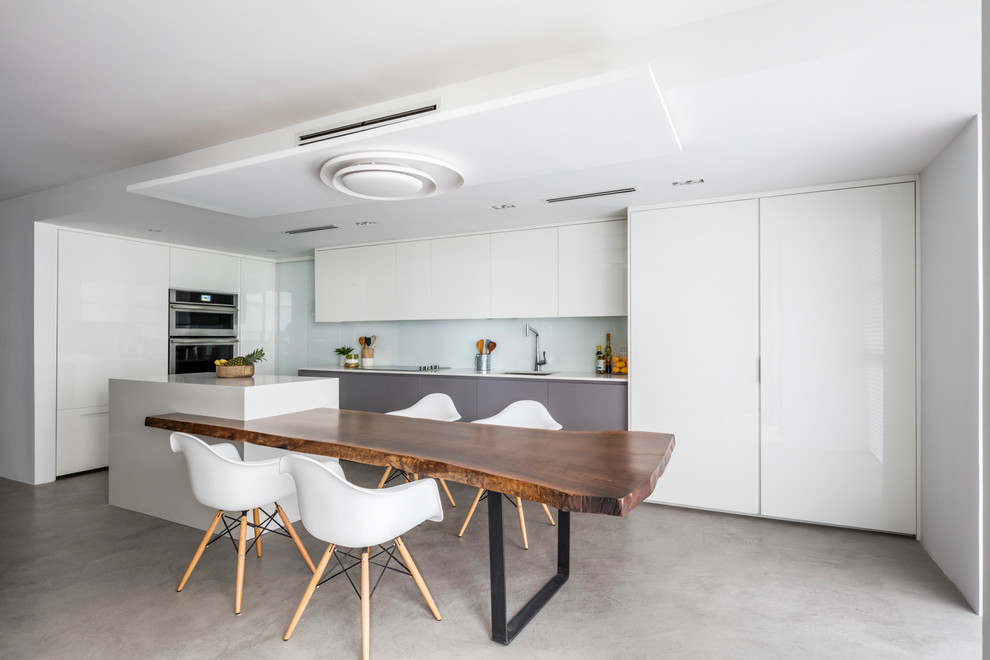 This is an example of a mid-sized contemporary l-shaped eat-in kitchen in Miami with an undermount sink, flat-panel cabinets, white cabinets, quartz benchtops, white splashback, glass sheet splashback, panelled appliances, concrete floors and with island.