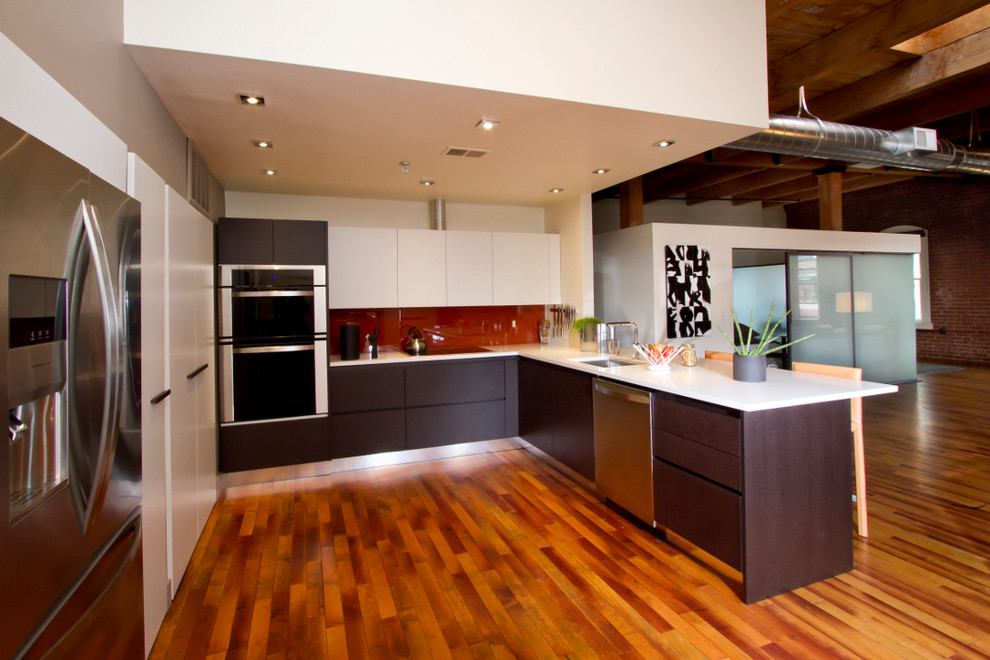 This is an example of an industrial u-shaped eat-in kitchen in Boston with an undermount sink, flat-panel cabinets, solid surface benchtops and stainless steel appliances.