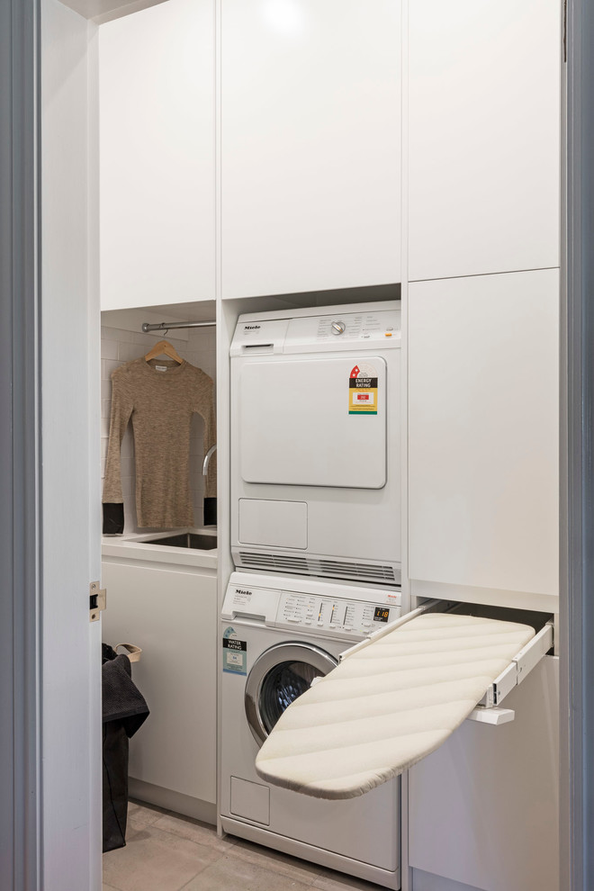 Photo of a mid-sized traditional galley dedicated laundry room in Sydney with recessed-panel cabinets, white cabinets, quartz benchtops, white walls, ceramic floors, a stacked washer and dryer and an undermount sink.