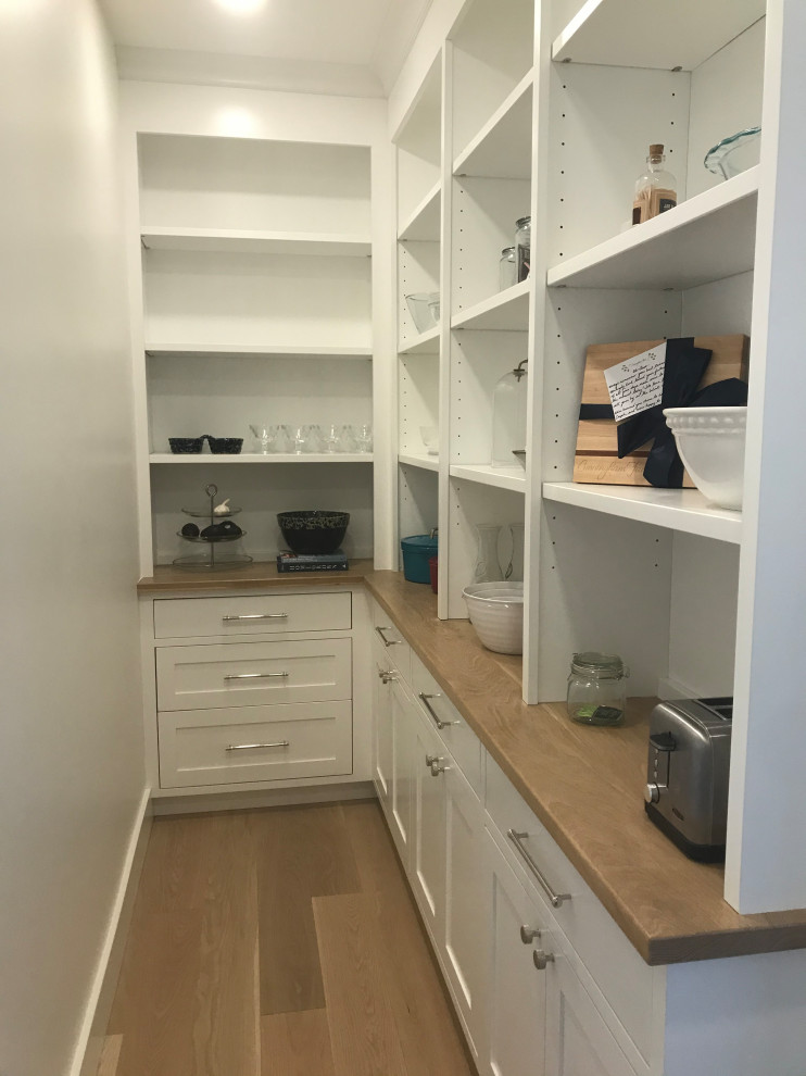 This is an example of a mid-sized traditional l-shaped kitchen pantry in Boston with a farmhouse sink, shaker cabinets, white cabinets, wood benchtops, stainless steel appliances, light hardwood floors, with island, brown floor and white benchtop.