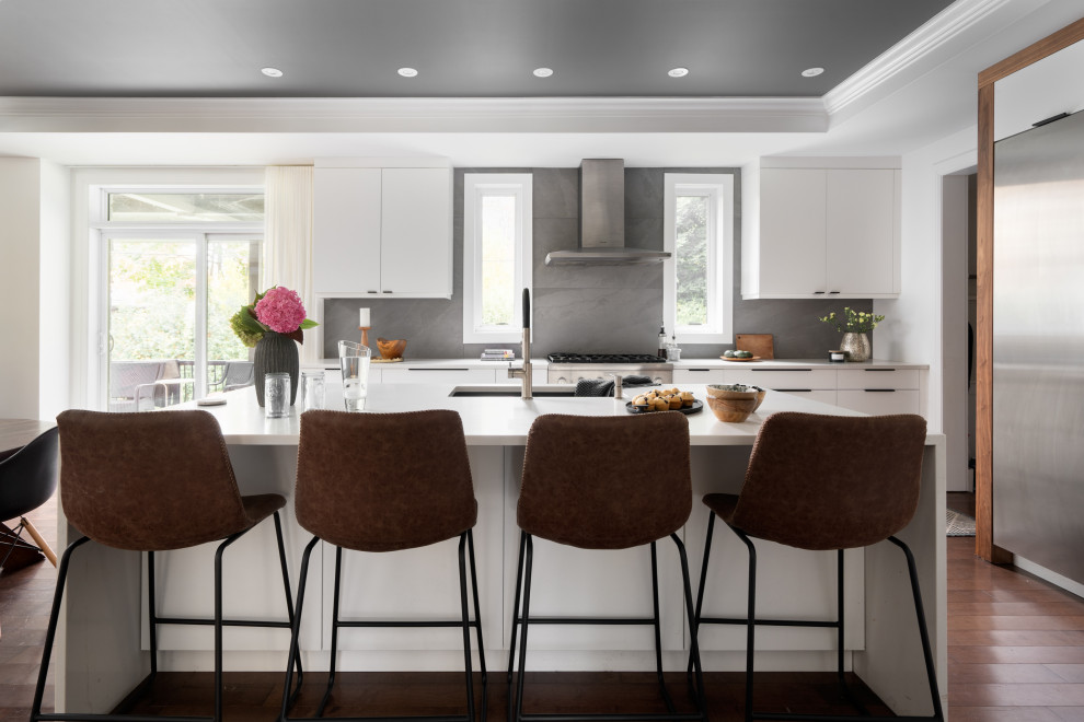 This is an example of a large transitional u-shaped eat-in kitchen in Montreal with an undermount sink, flat-panel cabinets, white cabinets, quartz benchtops, grey splashback, slate splashback, stainless steel appliances, dark hardwood floors, with island, brown floor, white benchtop and coffered.