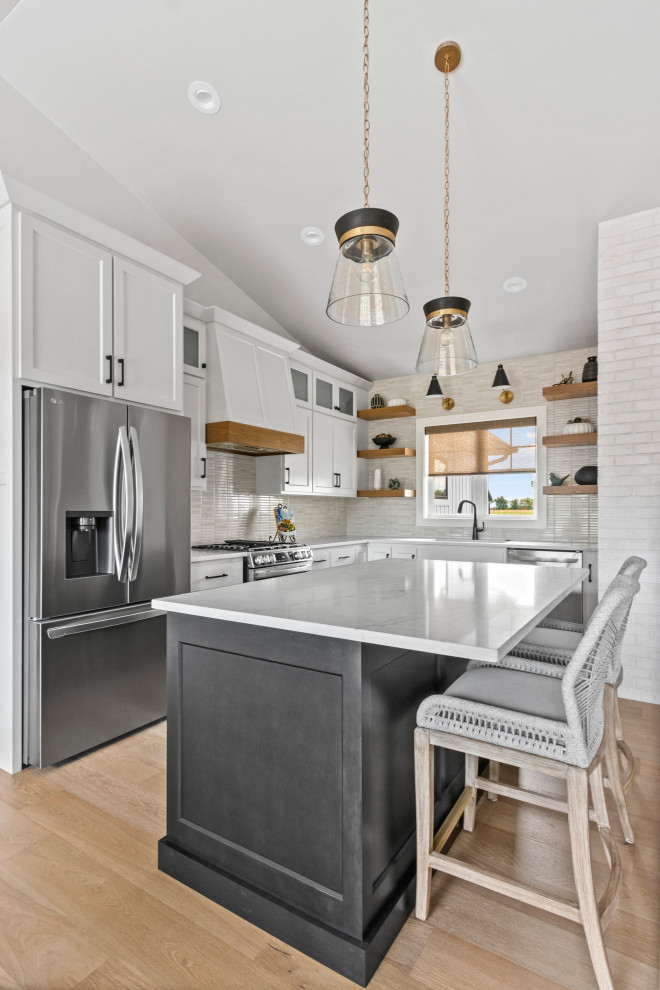 Example of a mid-sized transitional l-shaped medium tone wood floor, brown floor and vaulted ceiling eat-in kitchen design with an undermount sink, shaker cabinets, white cabinets, quartz countertops, white backsplash, porcelain backsplash, stainless steel appliances, an island and white countertops