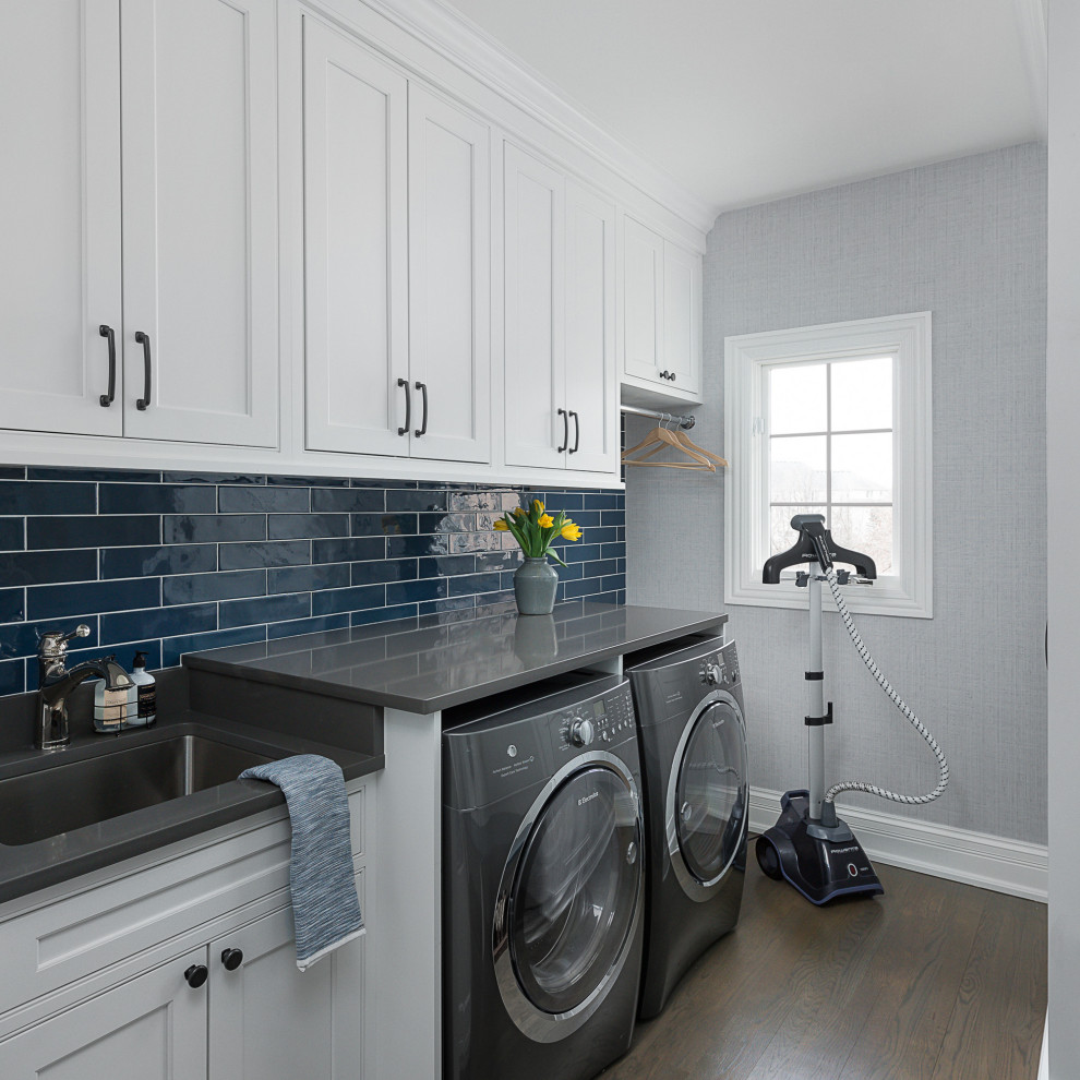 Design ideas for a mid-sized transitional galley utility room in Chicago with an undermount sink, beaded inset cabinets, white cabinets, quartz benchtops, white splashback, porcelain splashback, grey walls, medium hardwood floors, a side-by-side washer and dryer, brown floor and grey benchtop.