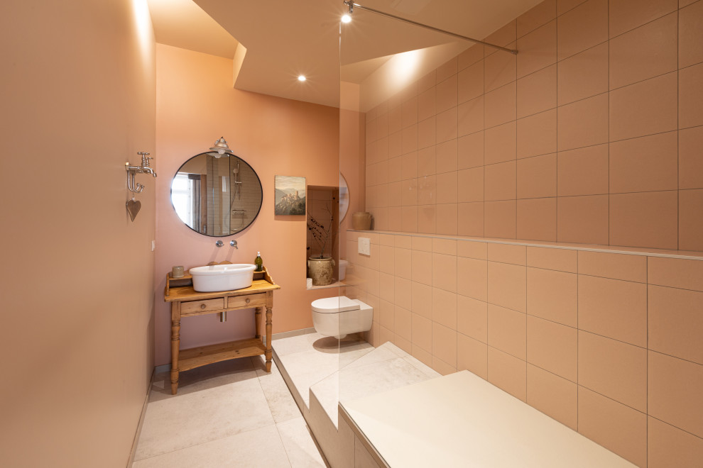 This is an example of a mid-sized traditional master bathroom in Strasbourg with open cabinets, medium wood cabinets, a curbless shower, a wall-mount toilet, pink tile, ceramic tile, pink walls, ceramic floors, a vessel sink, wood benchtops, beige floor, an open shower, brown benchtops, a niche, a single vanity, a freestanding vanity and recessed.
