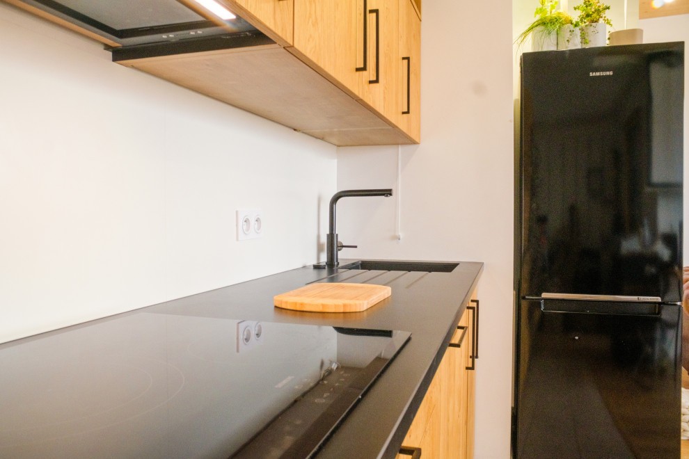 Идея дизайна: прямая кухня-гостиная среднего размера в стиле модернизм с светлыми деревянными фасадами, черной техникой, черной столешницей и монолитной мойкой без острова