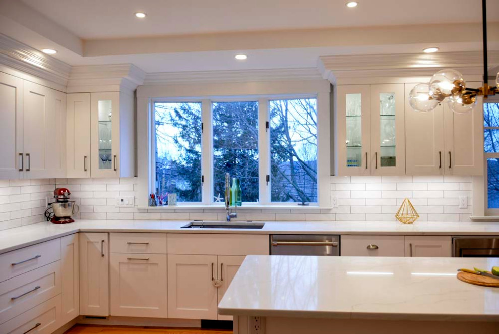 Large transitional l-shaped eat-in kitchen in Boston with an undermount sink, shaker cabinets, white cabinets, quartz benchtops, white splashback, ceramic splashback, stainless steel appliances, light hardwood floors, with island, brown floor and white benchtop.