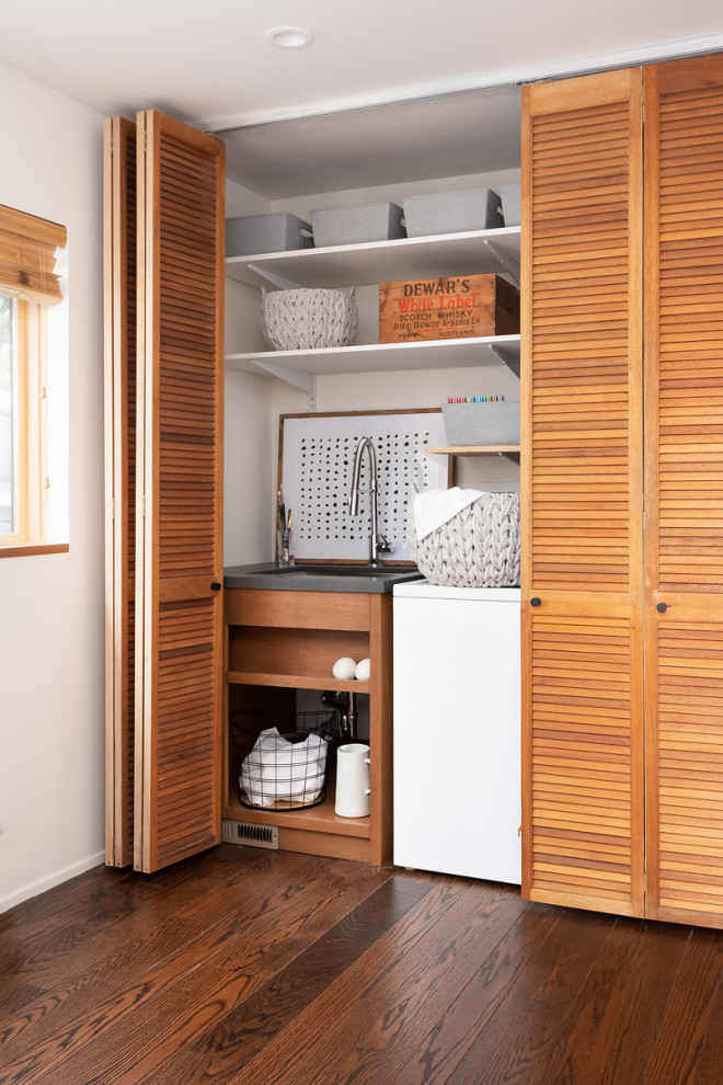 Large trendy medium tone wood floor and brown floor utility room photo in Sacramento with a single-bowl sink, brown cabinets, concrete countertops, white walls, a concealed washer/dryer and gray countertops