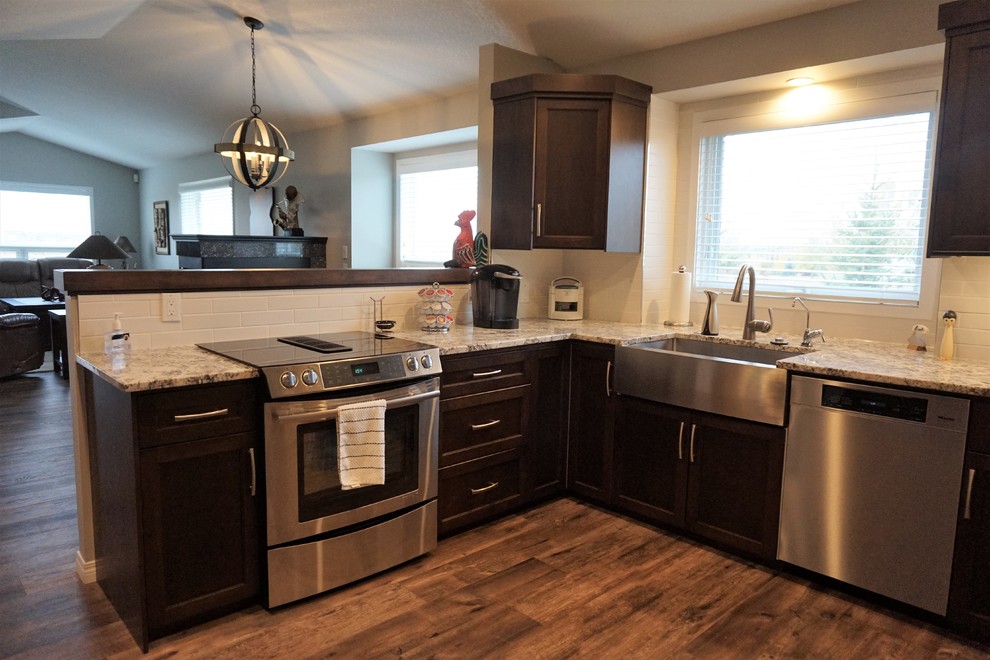 Example of a mid-sized transitional u-shaped dark wood floor and brown floor kitchen design in Calgary with a farmhouse sink, shaker cabinets, dark wood cabinets, granite countertops, white backsplash, subway tile backsplash, stainless steel appliances and a peninsula