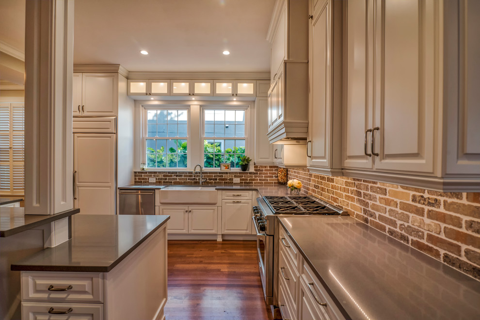 Photo of a large transitional l-shaped eat-in kitchen in Tampa with a farmhouse sink, raised-panel cabinets, white cabinets, limestone benchtops, red splashback, brick splashback, stainless steel appliances, dark hardwood floors, with island, brown floor and grey benchtop.
