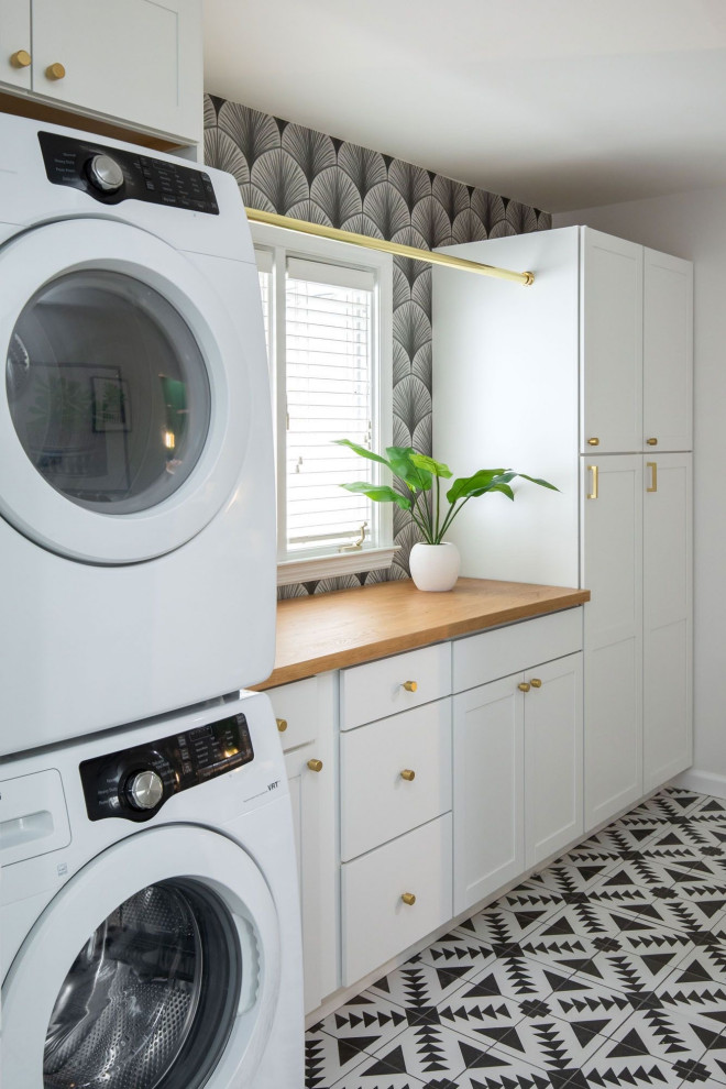Inspiration for a mid-sized transitional single-wall dedicated laundry room in Indianapolis with shaker cabinets, white cabinets, wood benchtops, white walls, ceramic floors, a stacked washer and dryer, black floor, brown benchtop and wallpaper.