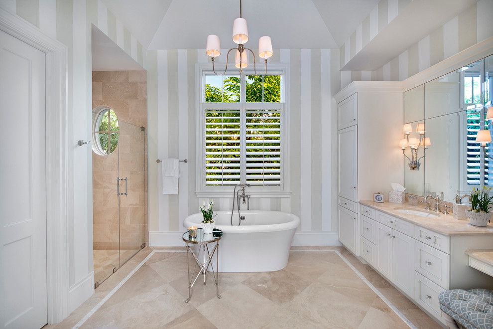 Photo of a large tropical master bathroom in Miami with shaker cabinets, white cabinets, a freestanding tub, an alcove shower, a two-piece toilet, beige tile, stone tile, multi-coloured walls, marble floors, an undermount sink and marble benchtops.