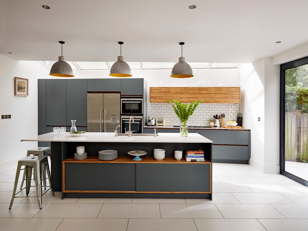 Large contemporary kitchen in London with flat-panel cabinets, grey cabinets, solid surface benchtops, white splashback, stainless steel appliances, with island and subway tile splashback.