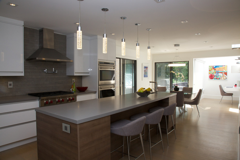 This is an example of a large modern single-wall eat-in kitchen in Toronto with a drop-in sink, flat-panel cabinets, grey cabinets, grey splashback, stainless steel appliances, medium hardwood floors, with island, brown floor, solid surface benchtops and stone tile splashback.