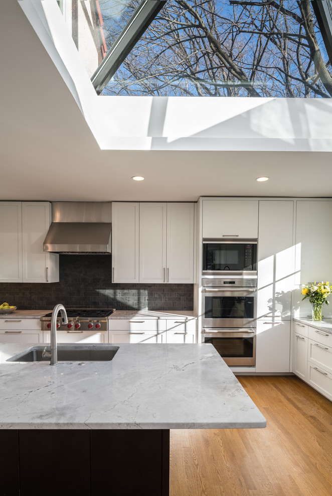 This is an example of a large contemporary u-shaped separate kitchen in DC Metro with a drop-in sink, recessed-panel cabinets, white cabinets, black splashback, slate splashback, stainless steel appliances, light hardwood floors, with island, brown floor and quartzite benchtops.