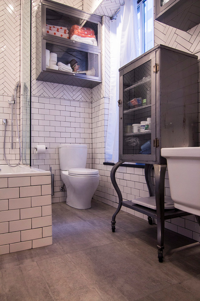 This is an example of a mid-sized industrial master bathroom in New York with a wall-mount sink, glass-front cabinets, grey cabinets, a drop-in tub, a corner shower, white tile, white walls, porcelain floors, brown floor, a one-piece toilet and subway tile.