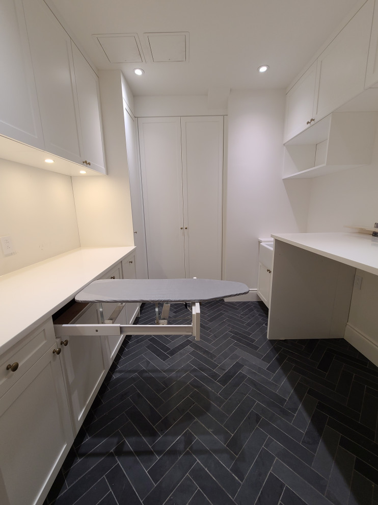 Photo of a large transitional galley dedicated laundry room in Montreal with a farmhouse sink, shaker cabinets, white cabinets, quartz benchtops, white splashback, white walls, slate floors, a side-by-side washer and dryer, grey floor and white benchtop.