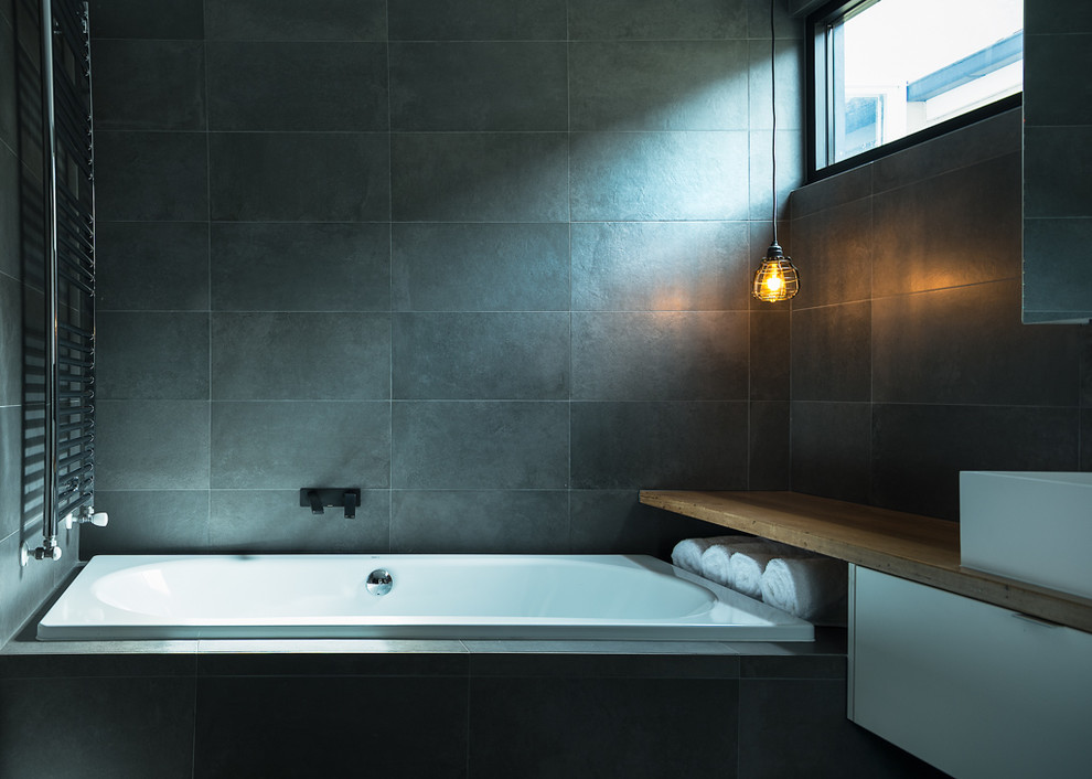 This is an example of a mid-sized industrial master bathroom in Melbourne with a vessel sink, flat-panel cabinets, white cabinets, wood benchtops, a drop-in tub, black tile, porcelain tile, black walls and brown benchtops.