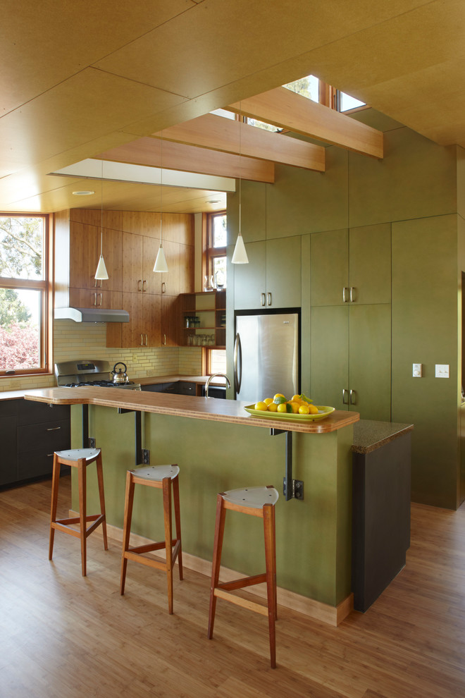 Mid-sized contemporary l-shaped open plan kitchen in San Francisco with flat-panel cabinets, green cabinets, green splashback, stainless steel appliances, medium hardwood floors, wood benchtops, glass tile splashback, with island and brown floor.