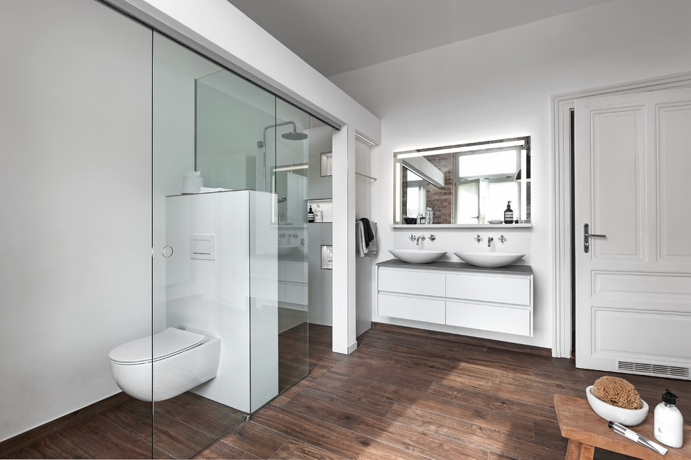 Inspiration for a large contemporary master bathroom in Frankfurt with flat-panel cabinets, grey cabinets, a freestanding tub, solid surface benchtops, brown floor, a sliding shower screen, medium hardwood floors, a curbless shower, a two-piece toilet, white walls, a vessel sink and white benchtops.