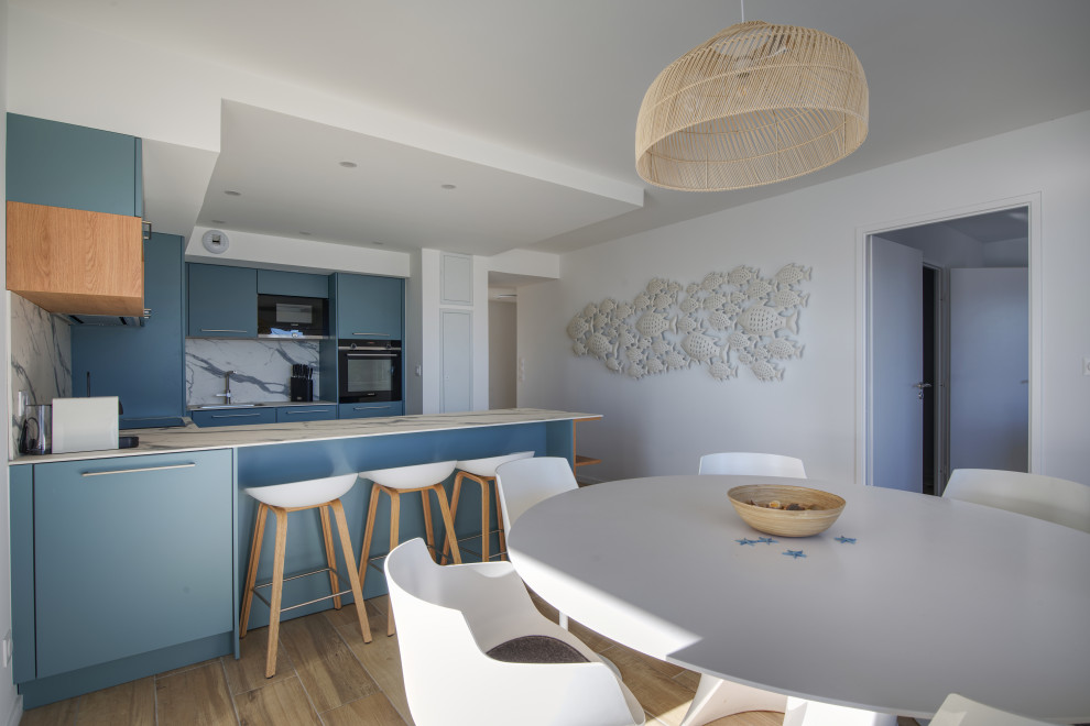This is an example of a mid-sized beach style u-shaped open plan kitchen in Montpellier with a single-bowl sink, blue cabinets, laminate benchtops, white splashback, panelled appliances, with island, white benchtop, light hardwood floors and beige floor.