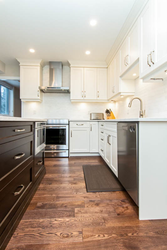 Example of a mid-sized transitional l-shaped medium tone wood floor and brown floor open concept kitchen design with an undermount sink, shaker cabinets, white cabinets, quartzite countertops, white backsplash, stone tile backsplash, stainless steel appliances, an island and white countertops