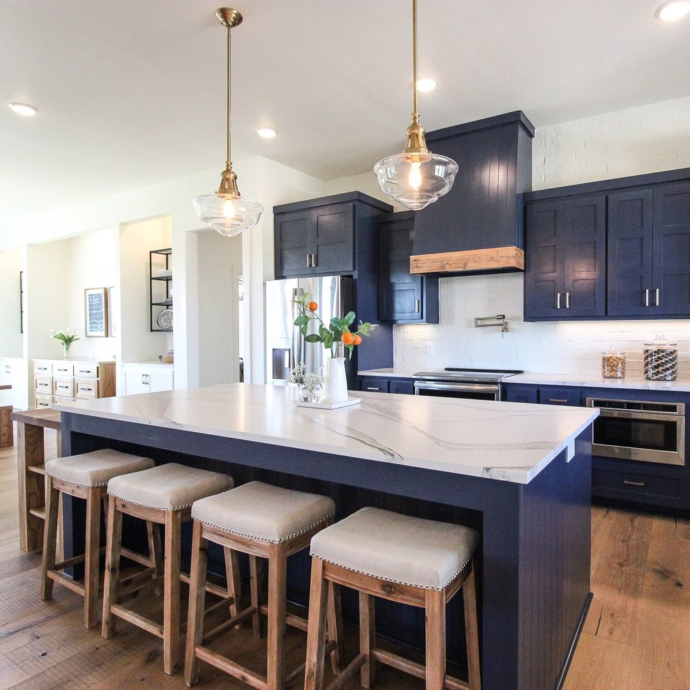 Design ideas for a transitional l-shaped eat-in kitchen in Dallas with a farmhouse sink, blue cabinets, quartz benchtops, white splashback, brick splashback, stainless steel appliances, light hardwood floors, with island and white benchtop.