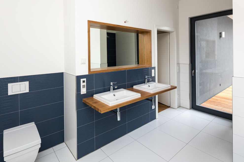 Photo of a large contemporary bathroom in Leipzig with a drop-in sink, wood benchtops, blue tile, white walls, a wall-mount toilet and ceramic floors.