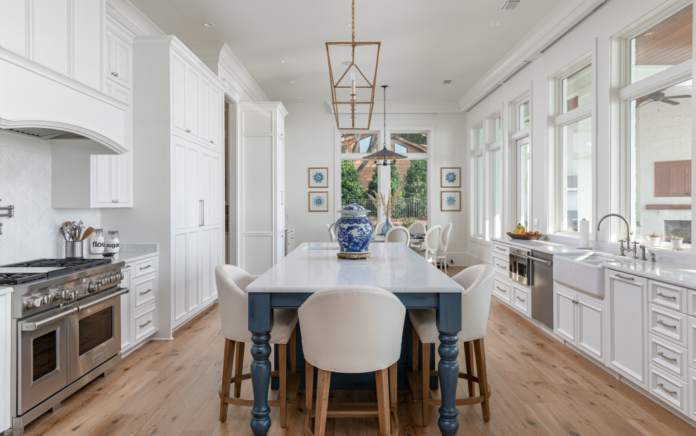Inspiration for a large contemporary u-shaped open plan kitchen in New Orleans with a farmhouse sink, beaded inset cabinets, white cabinets, white splashback, stainless steel appliances, light hardwood floors, with island, brown floor and white benchtop.