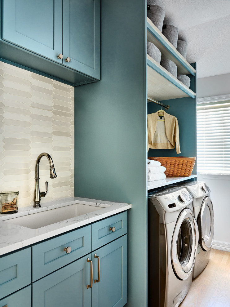 デンバーにあるお手頃価格の中くらいなおしゃれな洗濯室 (ll型、アンダーカウンターシンク、シェーカースタイル扉のキャビネット、青いキャビネット、人工大理石カウンター、ベージュキッチンパネル、セラミックタイルのキッチンパネル、白い壁、淡色無垢フローリング、左右配置の洗濯機・乾燥機、茶色い床、白いキッチンカウンター) の写真