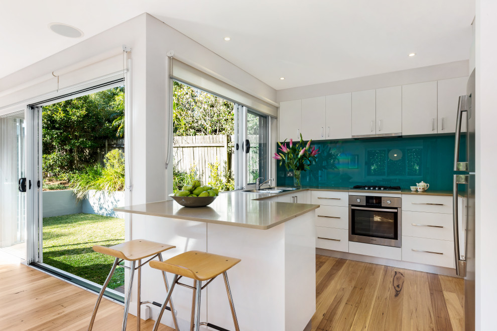This is an example of a contemporary u-shaped kitchen in Sunshine Coast with flat-panel cabinets, white cabinets, blue splashback, glass sheet splashback, stainless steel appliances, medium hardwood floors, a peninsula, brown floor and beige benchtop.