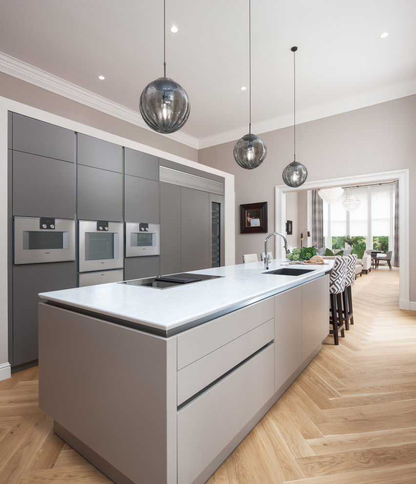 This is an example of a mid-sized contemporary galley kitchen in Glasgow with an undermount sink, flat-panel cabinets, grey cabinets, stainless steel appliances, light hardwood floors, with island, beige floor and white benchtop.