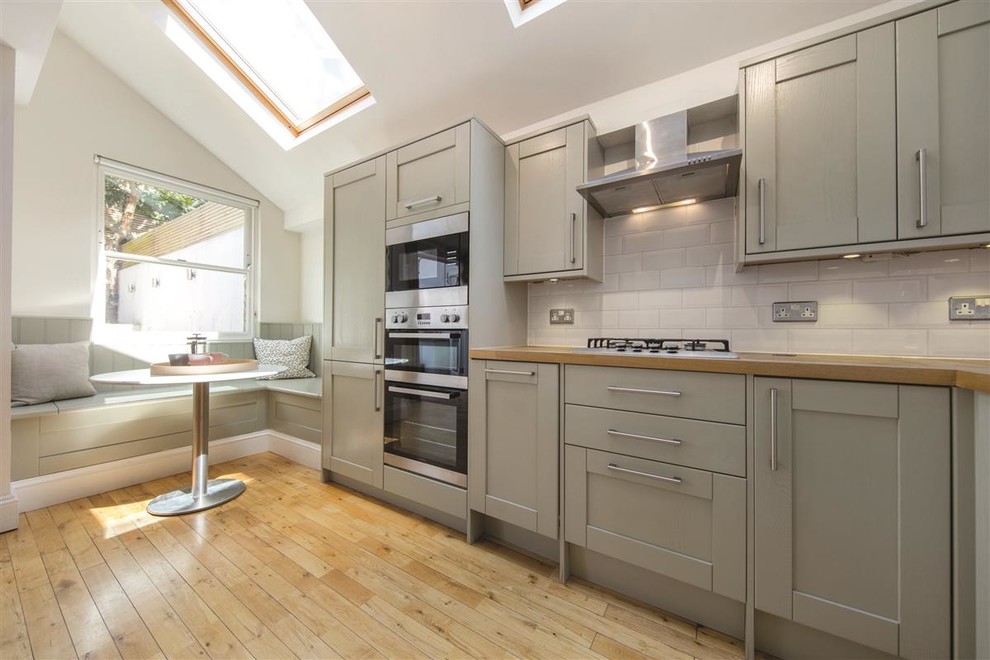 Photo of a small contemporary l-shaped kitchen in London with grey cabinets, wood benchtops, white splashback, stainless steel appliances, light hardwood floors, no island, beige floor, beige benchtop and recessed-panel cabinets.