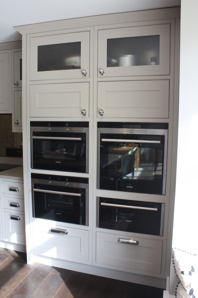 This is an example of a large classic l-shaped kitchen/diner in Hertfordshire with a submerged sink, shaker cabinets, beige cabinets, engineered stone countertops, metallic splashback, glass sheet splashback, stainless steel appliances, dark hardwood flooring and an island.