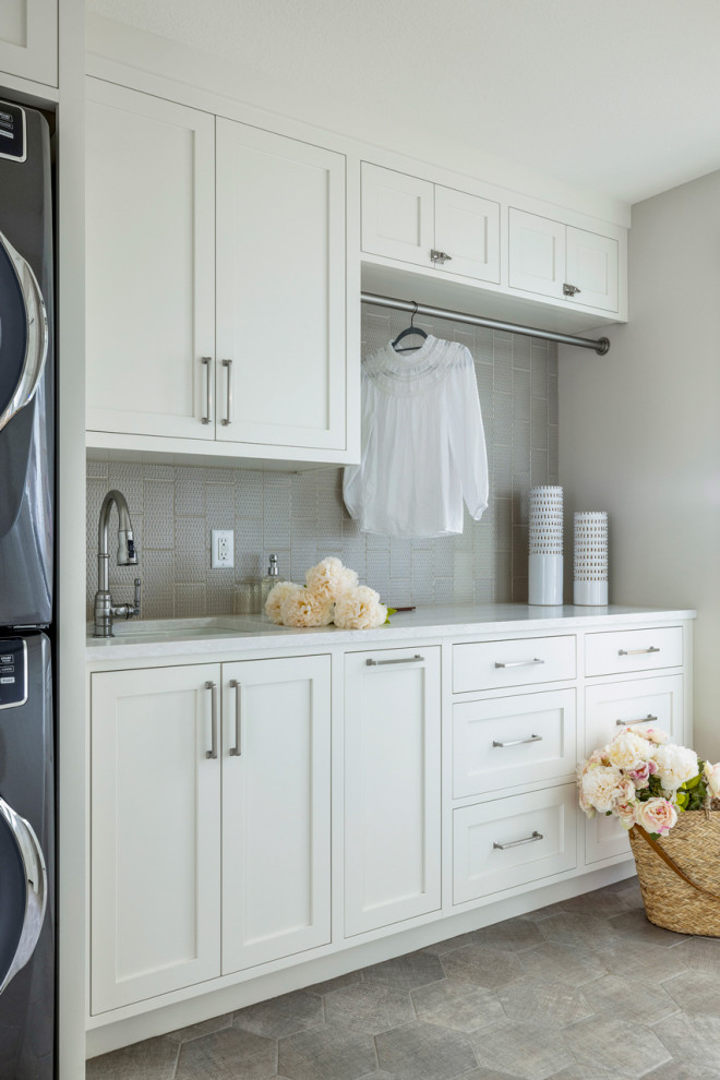 Design ideas for a small beach style galley dedicated laundry room in Minneapolis with a double-bowl sink, shaker cabinets, white cabinets, quartz benchtops, grey splashback, subway tile splashback, grey walls, porcelain floors, a stacked washer and dryer, grey floor and white benchtop.