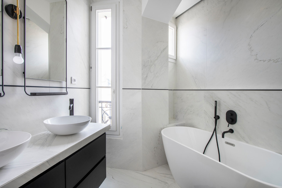 Inredning av ett modernt stort vit vitt en-suite badrum, med släta luckor, svarta skåp, ett fristående badkar, en kantlös dusch, en vägghängd toalettstol, vit kakel, marmorkakel, vita väggar, marmorgolv, ett fristående handfat, marmorbänkskiva, vitt golv och dusch med skjutdörr