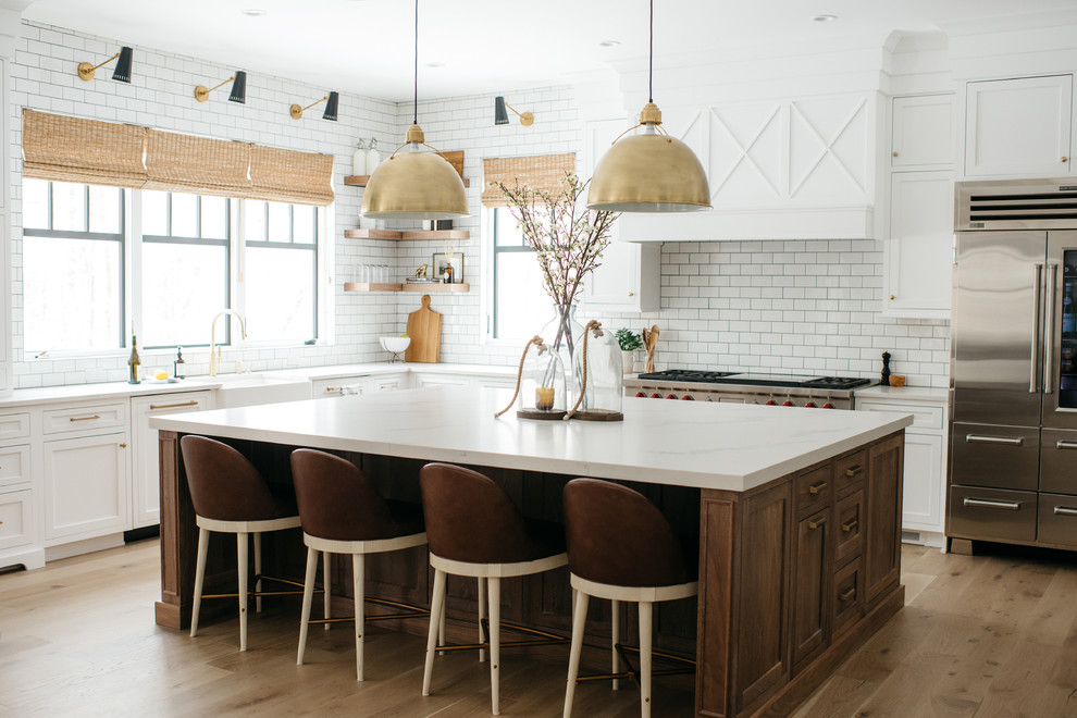 Country l-shaped eat-in kitchen in Detroit with a farmhouse sink, shaker cabinets, white cabinets, white splashback, subway tile splashback, stainless steel appliances, medium hardwood floors, with island, brown floor and white benchtop.