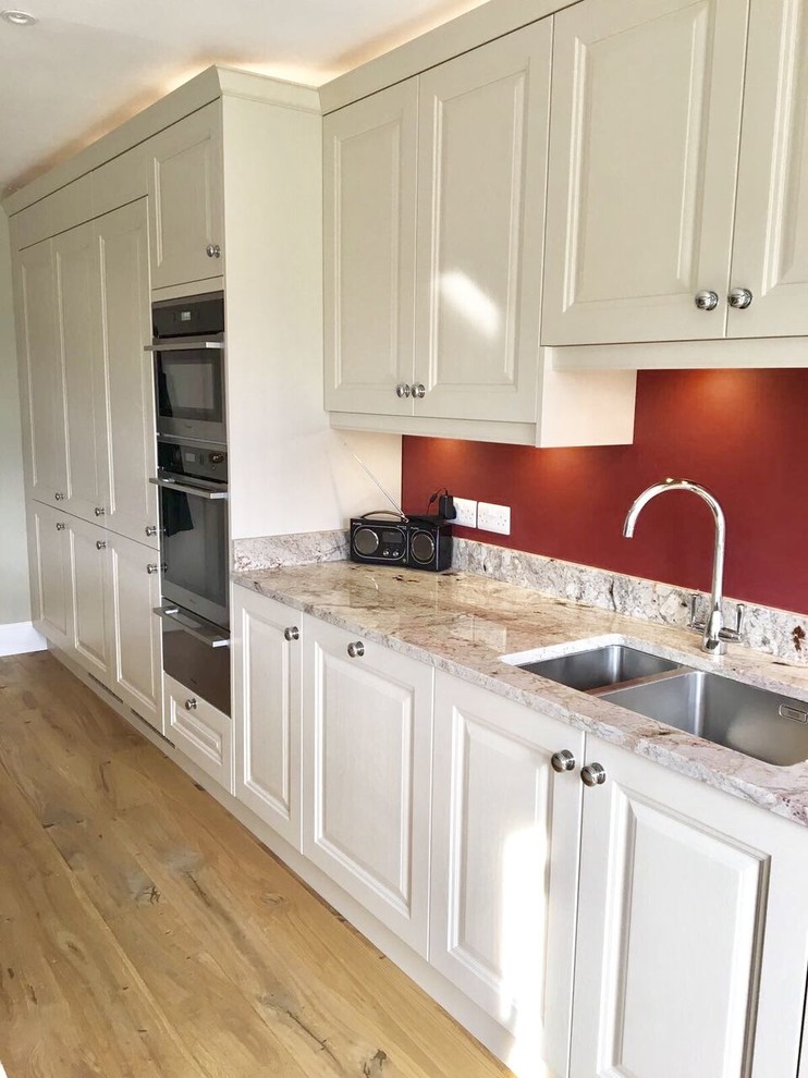 Large traditional open plan kitchen in Surrey with shaker cabinets, white cabinets, granite benchtops, panelled appliances, light hardwood floors and with island.