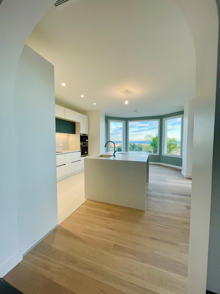 This is an example of a large contemporary l-shaped eat-in kitchen in Nice with an integrated sink, flat-panel cabinets, white cabinets, white splashback, ceramic splashback, panelled appliances, light hardwood floors, with island and white benchtop.