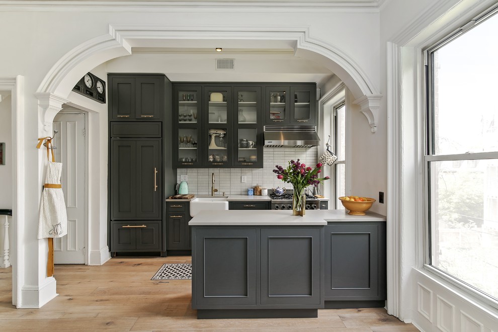 Mid-sized traditional galley eat-in kitchen in Philadelphia with a farmhouse sink, recessed-panel cabinets, grey cabinets, white splashback, medium hardwood floors, with island, beige floor, solid surface benchtops and ceramic splashback.