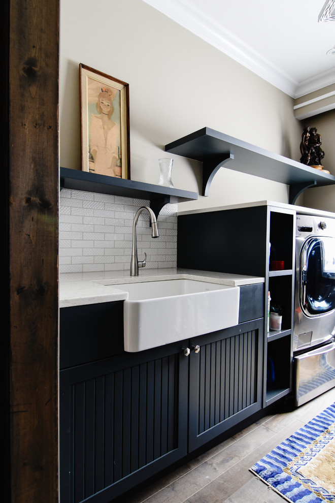 Design ideas for a mid-sized country galley dedicated laundry room in Montreal with a farmhouse sink, quartz benchtops, beige walls, medium hardwood floors, a side-by-side washer and dryer, recessed-panel cabinets, black cabinets and beige floor.