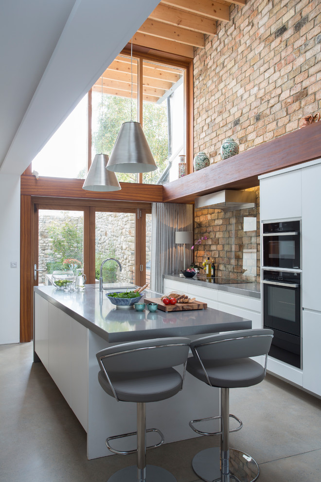 This is an example of a mid-sized contemporary kitchen in Other with flat-panel cabinets, white cabinets, black appliances, with island, grey floor, brick splashback and concrete floors.