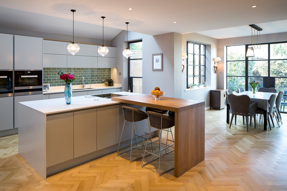 Photo of a contemporary galley kitchen in Hertfordshire with an undermount sink, flat-panel cabinets, white cabinets, green splashback, subway tile splashback, stainless steel appliances, medium hardwood floors, with island, brown floor and white benchtop.