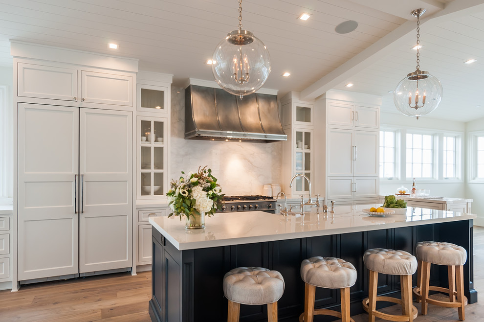 This is an example of a large country u-shaped open plan kitchen in Los Angeles with a farmhouse sink, recessed-panel cabinets, white cabinets, solid surface benchtops, white splashback, marble splashback, panelled appliances, light hardwood floors, with island and white benchtop.
